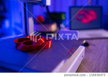 A red object is being printed on a 3D printer with a laptop displaying its design in the background, illuminated by blue light 138836473