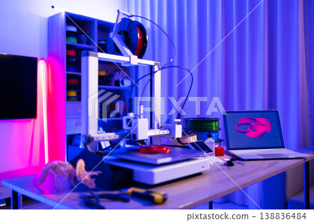 A 3D printer is actively creating an object while a laptop displays its design, surrounded by filament spools and tools under vibrant neon lighting A 3D printer is actively creating an object while a laptop displays its design, surrounded by filament spools and tools under vibrant neon lighting 138836484