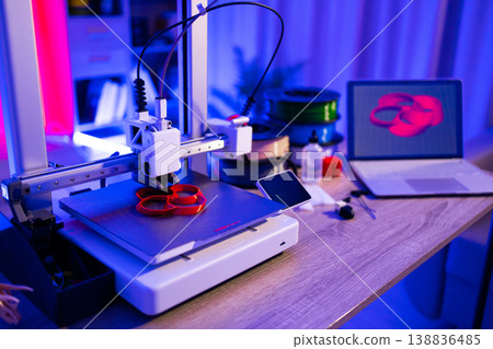 A 3D printer is actively creating a red object on its build plate, with filament spools and a laptop displaying a 3D model in the background 138836485