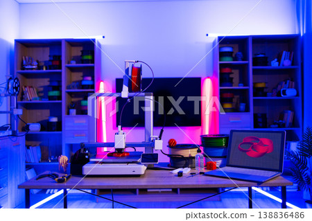 A modern 3D printer is set up on a desk with spools of filament and a laptop displaying a 3D model, illuminated by vibrant neon lighting 138836486