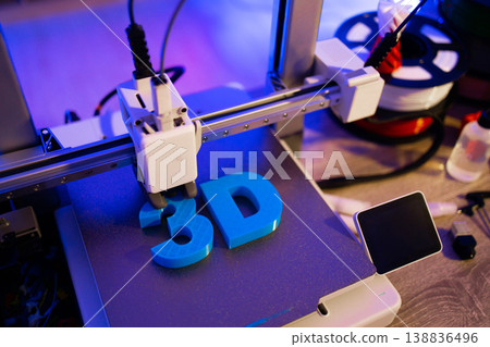 A 3D printer is actively creating blue letters on its build plate with filament spools in the background 138836496