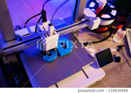 A close-up shot of a 3D printer in action, meticulously crafting blue letters on its build plate with vibrant purple lighting 138836498