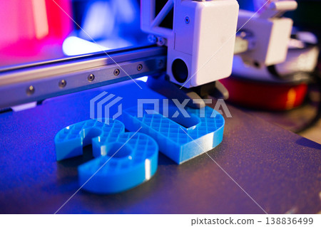 A 3D printer is actively creating blue plastic objects on its build plate with a close-up perspective 138836499