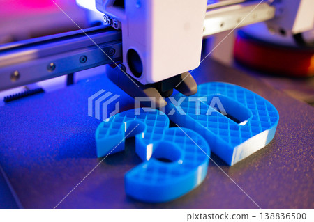 A close-up shot shows a 3D printer's nozzle depositing blue plastic to form the numbers 3 and 0 on a textured surface under purple lighting A close-up shot shows a 3D printer's nozzle depositing blue plastic to form the numbers 3 and 0 on a textured surface under purple lighting 138836500