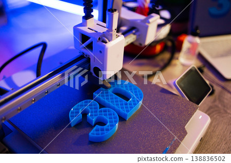 A modern 3D printer is actively creating bright blue letters on a textured build plate with vibrant blue and purple lighting 138836502