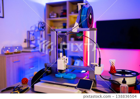 A modern 3D printer is actively creating blue objects on its platform with spools of filament nearby and colorful ambient lighting A modern 3D printer is actively creating blue objects on its platform with spools of filament nearby and colorful ambient lighting 138836504