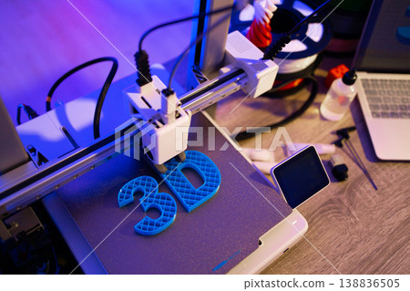 A 3D printer is actively creating blue letters on a textured surface, with filament spools and a laptop visible in the background under purple lighting A 3D printer is actively creating blue letters on a textured surface, with filament spools and a laptop visible in the background under purple lighting 138836505