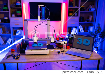 A 3D printer is actively creating a blue object on a desk in a room illuminated by vibrant neon lights, with a laptop displaying the 3D logo nearby A 3D printer is actively creating a blue object on a desk in a room illuminated by vibrant neon lights, with a laptop displaying the 3D logo nearby 138836515