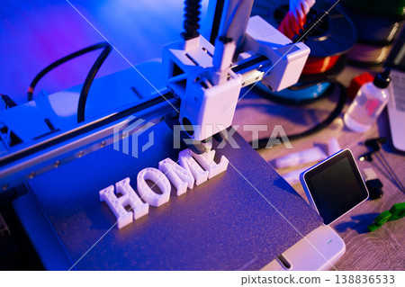 A 3D printer is actively creating the word HOME in white plastic letters on its build plate with vibrant blue and purple lighting A 3D printer is actively creating the word HOME in white plastic letters on its build plate with vibrant blue and purple lighting 138836533