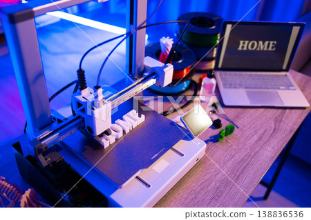 A 3D printer is actively creating the word HOME in white plastic filament on its build plate, surrounded by spools of filament and a laptop displaying the same word 138836536