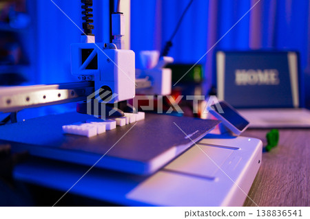 A modern 3D printer is actively creating small white objects on its build plate under blue lighting A modern 3D printer is actively creating small white objects on its build plate under blue lighting 138836541