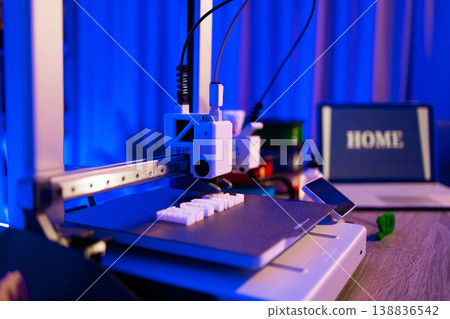 A 3D printer is actively creating white plastic objects on its build plate under vibrant blue lighting, with a laptop displaying the word HOME in the background 138836542
