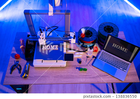 A 3D printer is actively creating the word HOME while filament spools and tools are laid out on a wooden desk 138836550