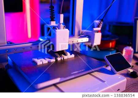 A modern 3D printer is actively creating white plastic letters on its build plate with vibrant neon lighting 138836554