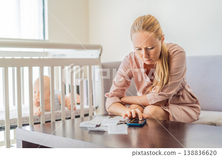 Mother sitting on sofa calculating monthly expenses with receipts while baby lies in crib nearby at home. Stressed mom managing budget and childcare. Financial pressure and parenthood concept Mother sitting on sofa calculating monthly expenses with receipts while baby lies in crib nearby at home. Stressed mom managing budget and childcare. Financial pressure and parenthood concept 138836620