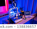 A 3D printer is actively creating the word HOME in white filament on its build plate, surrounded by spools of filament and a laptop displaying the same word 138836657