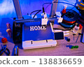 A 3D printer is actively creating the word HOME in white letters on its build plate, surrounded by filament spools and tools 138836659