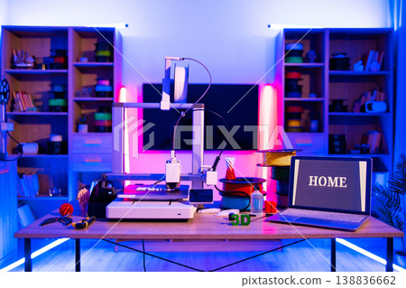 A 3D printer is set up on a desk with spools of filament and a laptop displaying the word HOME, illuminated by colorful LED lights 138836662