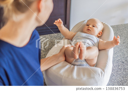 Mother talking to baby lying in cozy lounger on kitchen table while cooking at home. Caring mom interacting with infant during daily routine. Parenting, multitasking and family life concept Mother talking to baby lying in cozy lounger on kitchen table while cooking at home. Caring mom interacting with infant during daily routine. Parenting, multitasking and family life concept 138836753