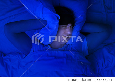 Frustrated man in a sleep mask covering ears with a pillow in bed at night. Concept of insomnia, noisy neighbors, and poor sleep causing irritation and stress. Frustrated man in a sleep mask covering ears with a pillow in bed at night. Concept of insomnia, noisy neighbors, and poor sleep causing irritation and stress. 138838223