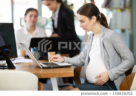 Pregnant woman is working on a laptop in an office while colleagues are discussing a project Pregnant woman is working on a laptop in an office while colleagues are discussing a project 138839202