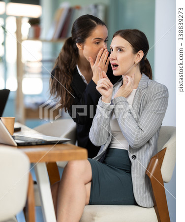 Female employee sharing gossip with surprised young colleague Female employee sharing gossip with surprised young colleague 138839273