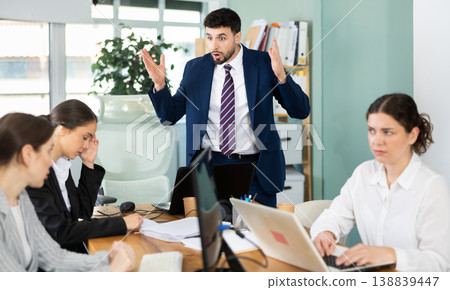 Young man standing and scolding sad seated colleagues in office Young man standing and scolding sad seated colleagues in office 138839447
