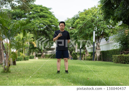Asian man wearing black sportswear stands on grass in a residential park using a smartphone during an outdoor fitness activity. Asian man wearing black sportswear stands on grass in a residential park using a smartphone during an outdoor fitness activity. 138840437
