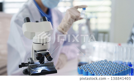 Adult researcher wearing mask examines sample vial with laboratory microscope Scientific discovery medical innovation healthcare technology development 138841403