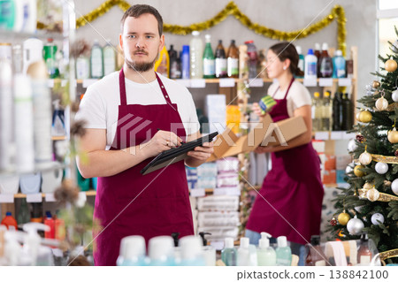 Focused bearded man seller restock of goods using tablet, female assistant laying out goods in background in store decorated for New Year holiday Focused bearded man seller restock of goods using tablet, female assistant laying out goods in background in store decorated for New Year holiday 138842100