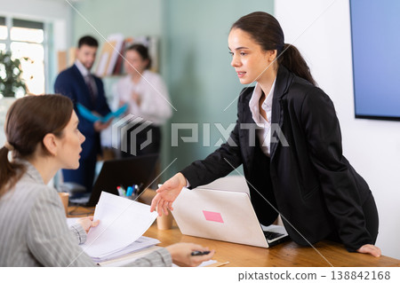 Female manager leaning over table and instructing young employee Female manager leaning over table and instructing young employee 138842168