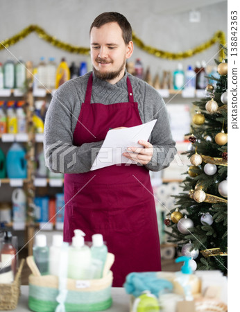 Attentive male salesperson restock of goods, checking papers in small household store decorated for New Year celebration 138842363