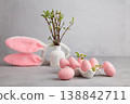 Pink Easter eggs on a tray next to a vase with green leafy branches and a bunny-ear headpiece.  138842711