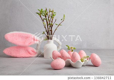 Pink Easter eggs on a tray next to a vase with green leafy branches and a bunny-ear headpiece.  138842711