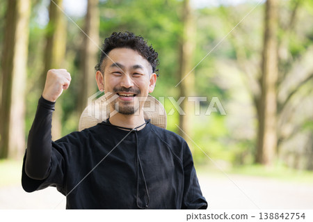 一位日本男子麵帶微笑,看著鏡頭,在綠樹成蔭的戶外露營地擺出勝利的姿勢,讓人聯想到戶外休閒和郊遊的景象。 一位日本男子麵帶微笑,看著鏡頭,在綠樹成蔭的戶外露營地擺出勝利的姿勢,讓人聯想到戶外休閒和郊遊的景象。 138842754