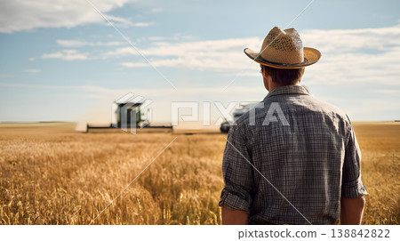 A adult white american farmer man standing on a wheat grass field. wearing a hat. photo taken from behind his back. rain harvester in the background, sunny day. Farmer standing in golden wheat field d 138842822