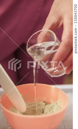 Pouring water powder. Cosmetic mask preparing. Unrecognizable beautician woman hands stirring facial mixture adding clear liquid from glass into bowl using spatula. Pouring water powder. Cosmetic mask preparing. Unrecognizable beautician woman hands stirring facial mixture adding clear liquid from glass into bowl using spatula. 138843700