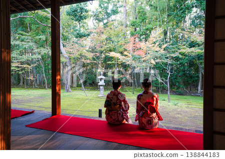 The backs of two women in kimonos sitting on a red coin and looking out at the garden. 138844183