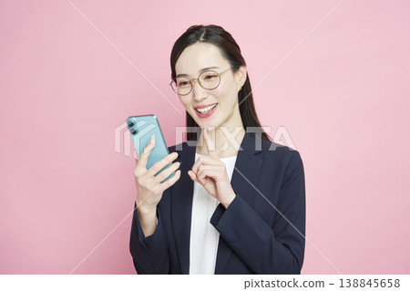 A smiling businesswoman looking at her smartphone against a pink background. A smiling businesswoman looking at her smartphone against a pink background. 138845658