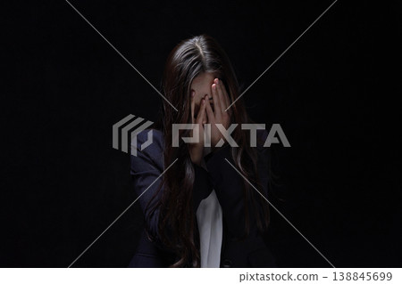 A businesswoman covers her face and looks dejected against a black background. A businesswoman covers her face and looks dejected against a black background. 138845699