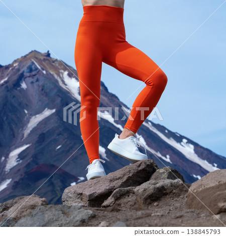 Athletic woman in orange leggings and white sneakers posing on rock with mountain view Athletic woman in orange leggings and white sneakers posing on rock with mountain view 138845793
