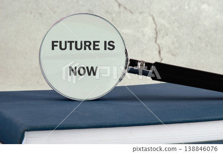 A magnifying glass magnifies the bold text Future is Now written on a dark blue book, emphasizing the importance of present actions 138846076