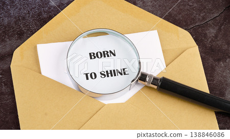 A magnifying glass rests on white paper, showing the phrase born to shine, peeking from a brown envelope on a dark textured background 138846086