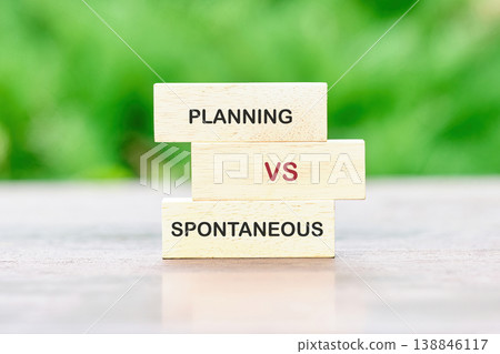 Stacked wooden blocks present PLANNING versus SPONTANEOUS on a light surface. Blurred green foliage in the background sets an outdoor scene Stacked wooden blocks present PLANNING versus SPONTANEOUS on a light surface. Blurred green foliage in the background sets an outdoor scene 138846117
