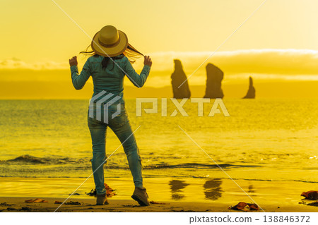 Woman in straw hat on sandy beach enjoying golden sunset over ocean with distant rock formations Woman in straw hat on sandy beach enjoying golden sunset over ocean with distant rock formations 138846372
