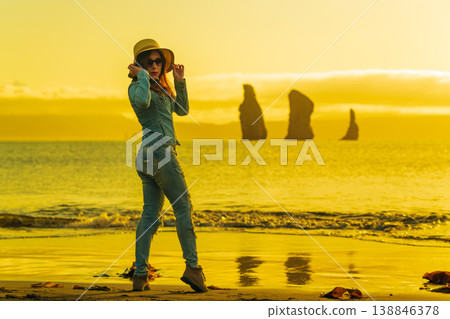 Stylish woman in hat and denim outfit on sandy beach during vibrant golden sunset 138846378