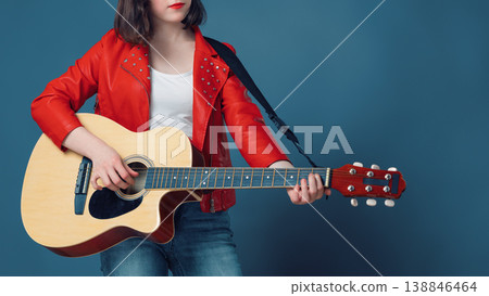 Stylish female musician in a red jacket playing an acoustic guitar against a blue studio background Stylish female musician in a red jacket playing an acoustic guitar against a blue studio background 138846464