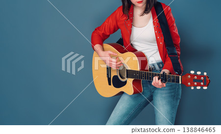 Female musician in red jacket and jeans playing an acoustic guitar against blue studio background Female musician in red jacket and jeans playing an acoustic guitar against blue studio background 138846465