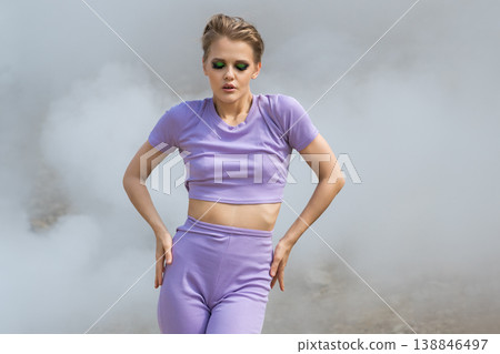 Stylish woman in lavender activewear with bold green eyeshadow posing in dramatic fog Stylish woman in lavender activewear with bold green eyeshadow posing in dramatic fog 138846497