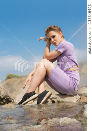 Woman with short hair in a lavender outfit posing on rocks by a natural hot spring under a blue sky Woman with short hair in a lavender outfit posing on rocks by a natural hot spring under a blue sky 138846498